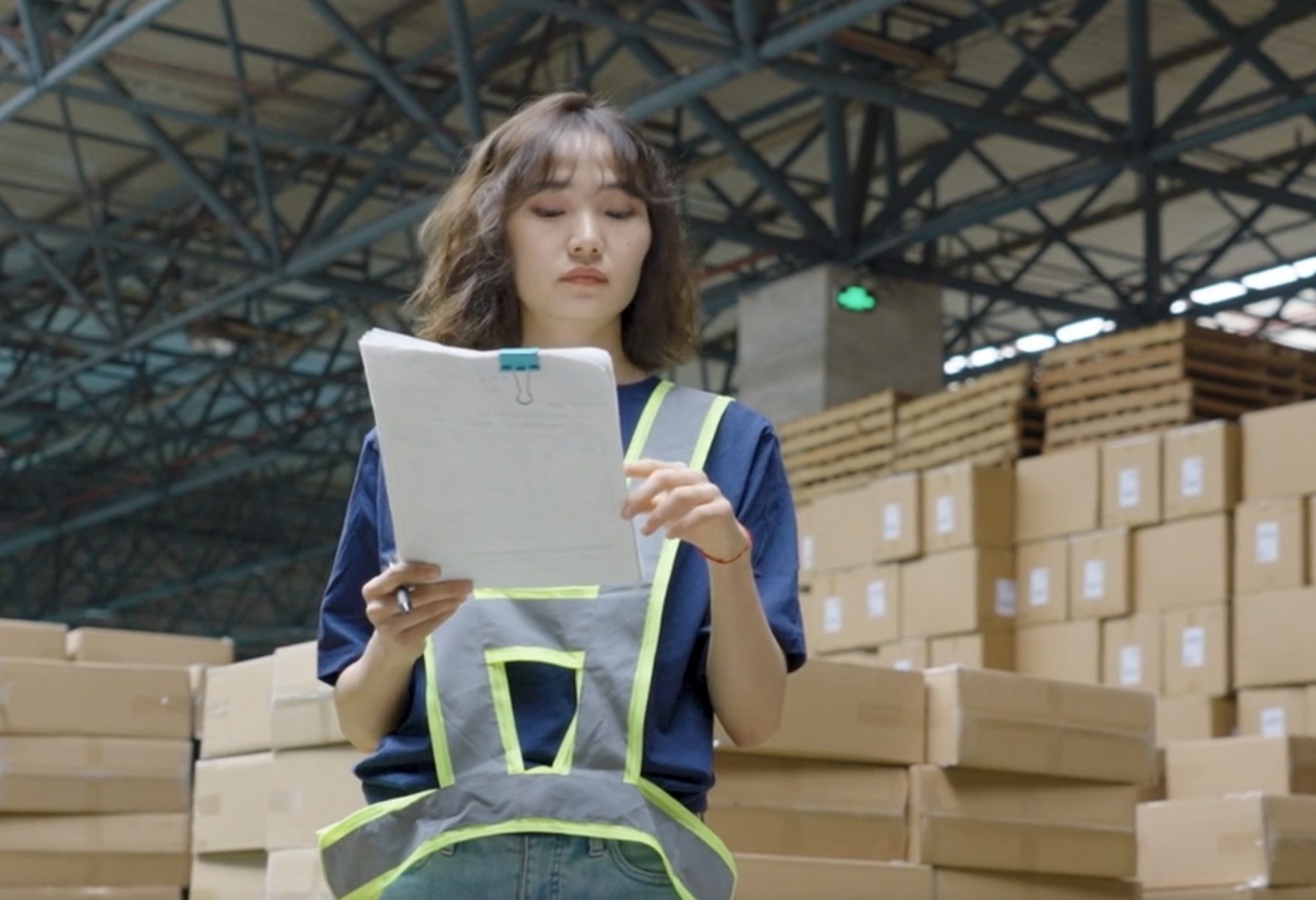 Warehouse operator in a hi-vis vest reviewing paperwork among stacked cartons